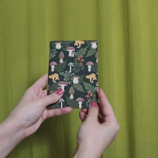 Video of two hands holding and flipping through a small sketchbook with mushroom pattern against a green curtain background