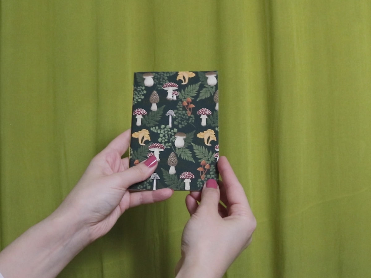 Video of two hands holding and flipping through a small sketchbook with mushroom pattern against a green curtain background
