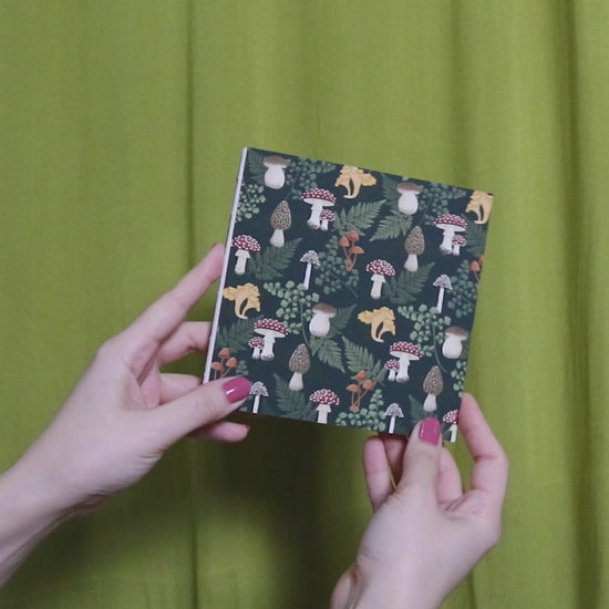 Video of two hands holding and flipping through a square sketchbook with mushroom pattern against a green curtain background