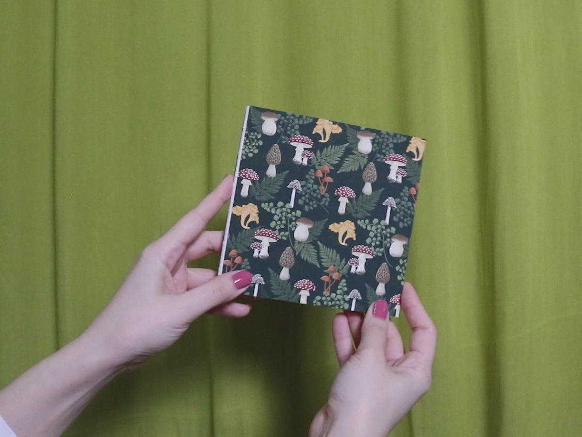 Video of two hands holding and flipping through a square sketchbook with mushroom pattern against a green curtain background