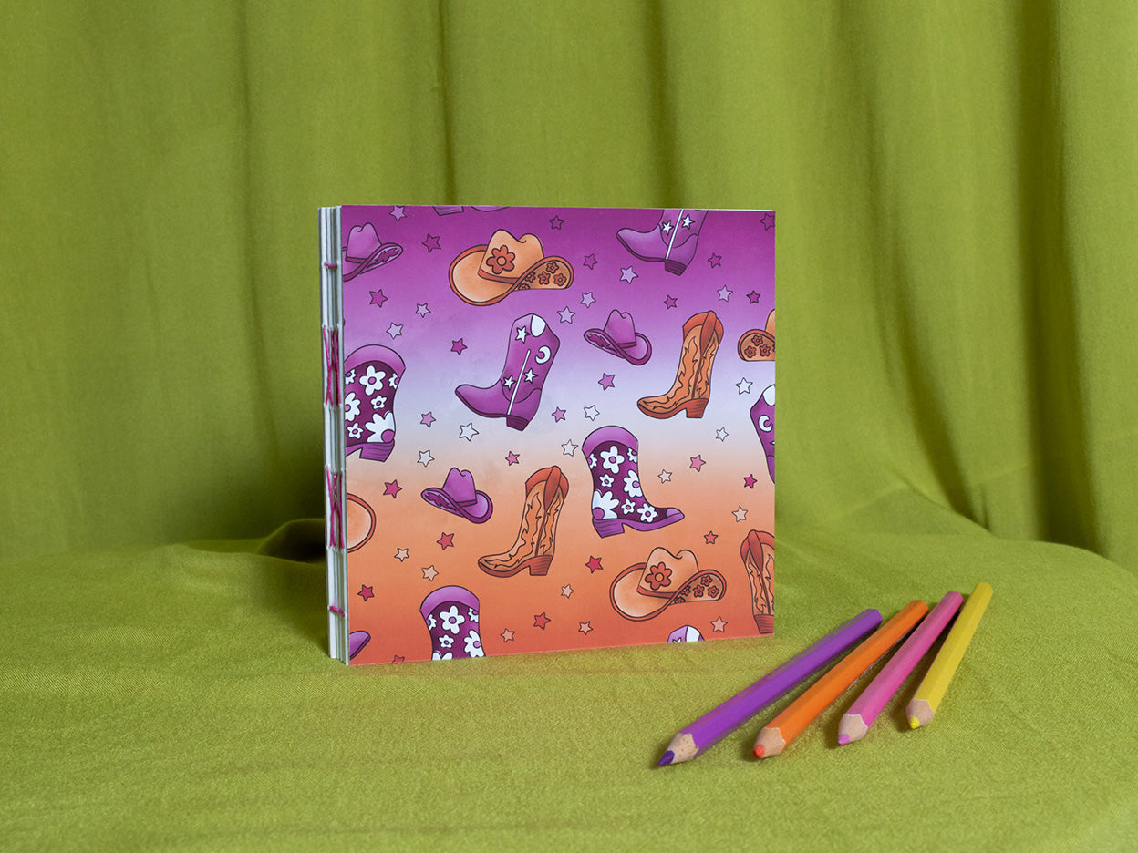 Square sketchbook with cowboy boot pattern on a green background with coloured pencils.