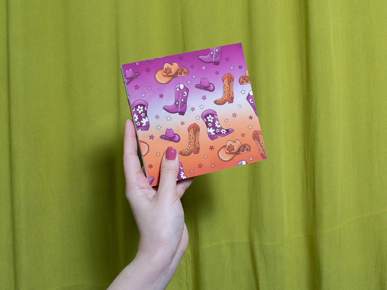 Hand holding a colourful square sketchbook with cowboy boot design against a green curtain background