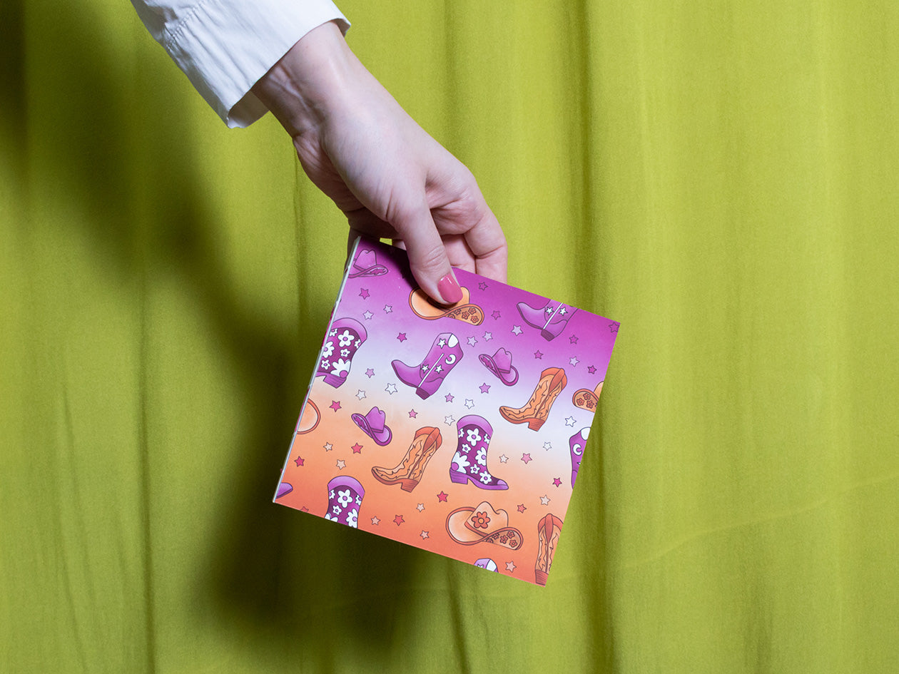 Hand holding a colourful square sketchbook with cowboy designs against a green background