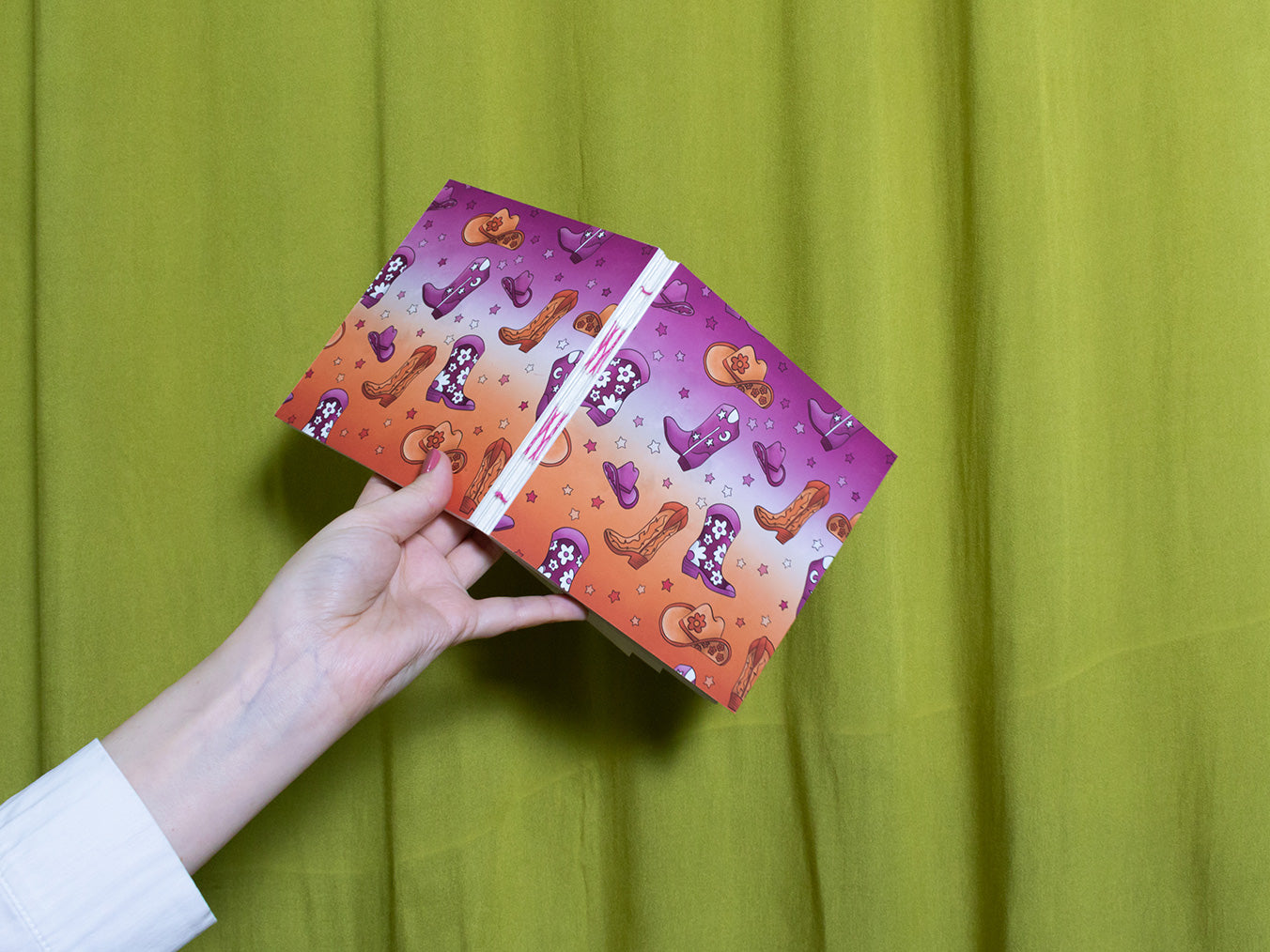 Hand holding a colourful square sketchbook with cowboy pattern against a green curtain background