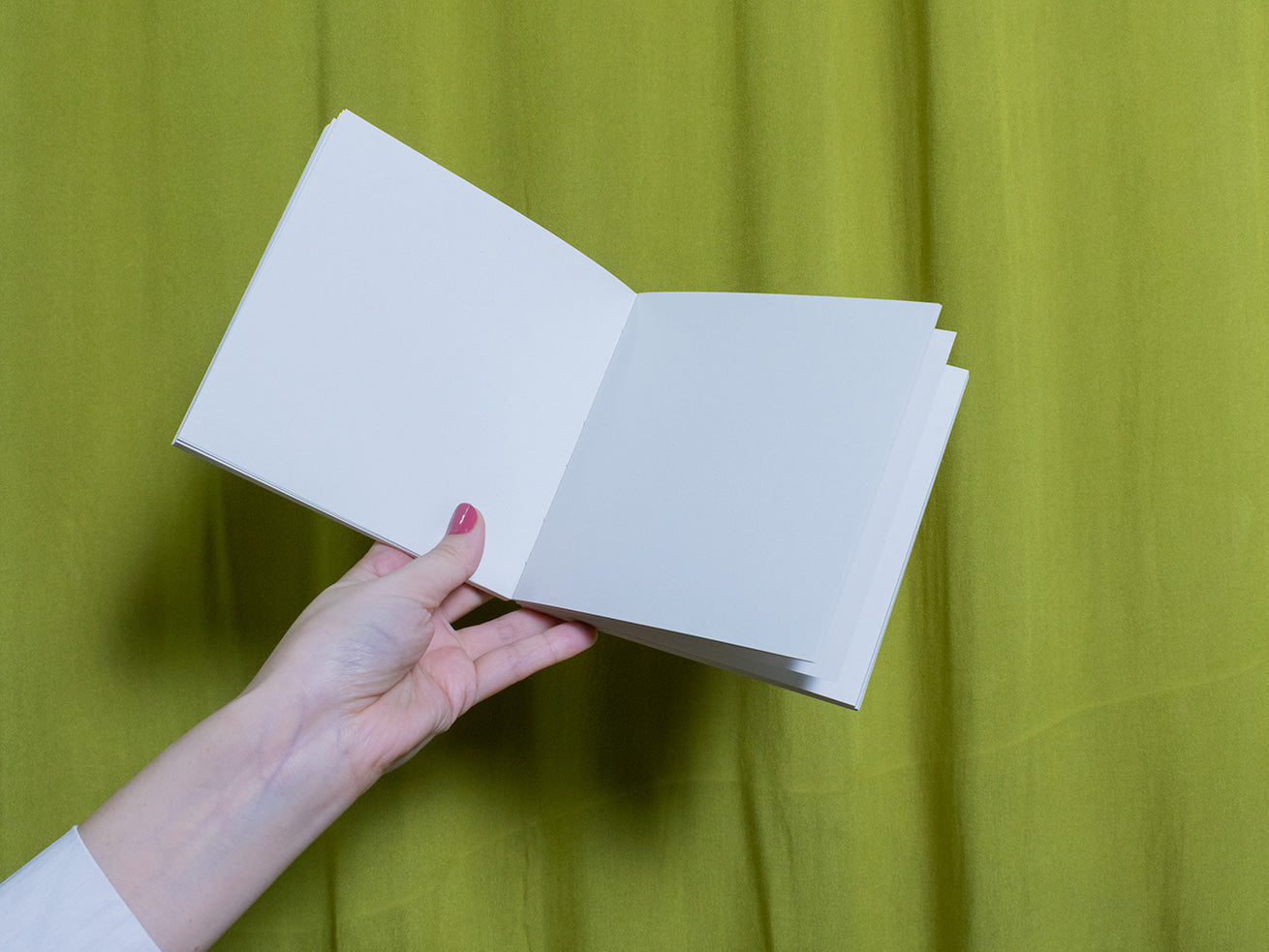 Hand holding a blank square sketchbook against a green curtain background
