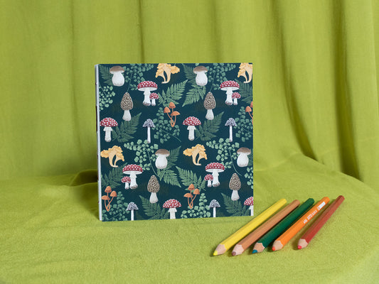 Square sketchbook with nature-themed mushroom cover and coloured pencils on a green fabric background