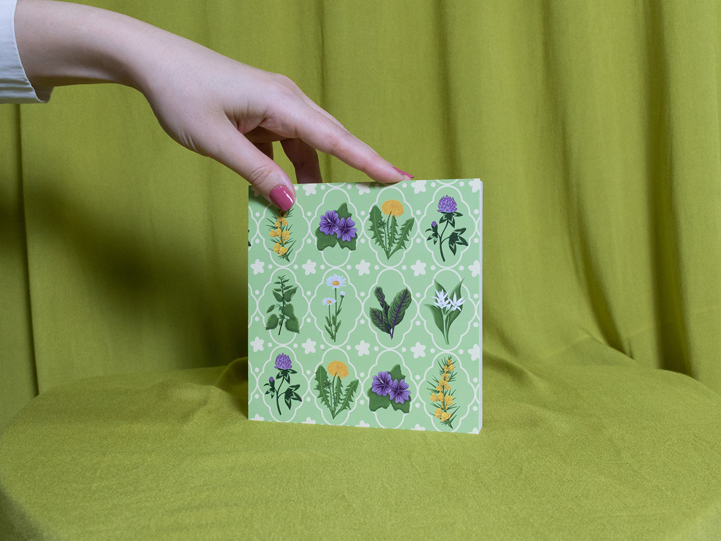 Hand holding a sketchbook with floral pattern against a green background