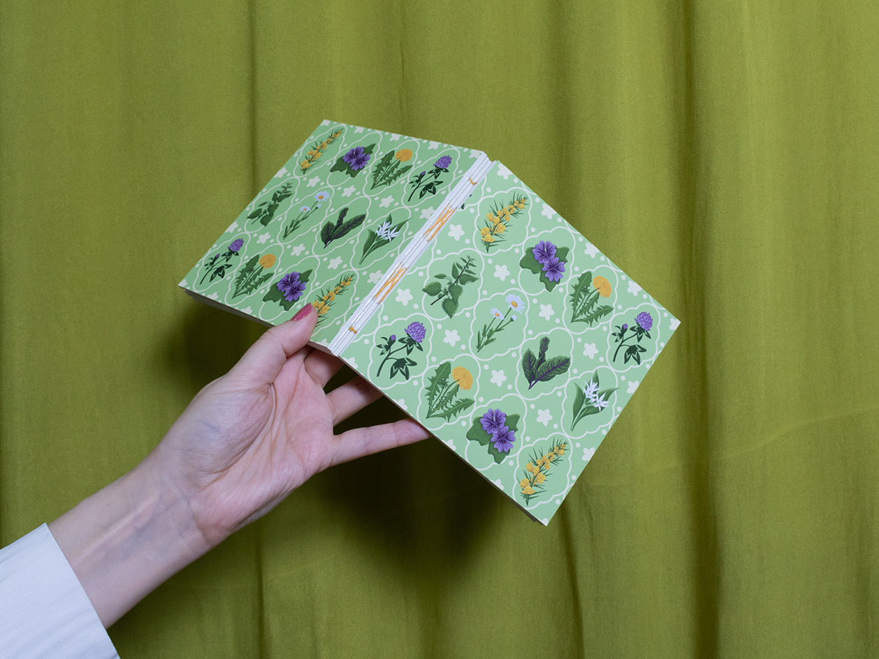 Hand holding a green sketchbook with floral patterns against a green curtain background