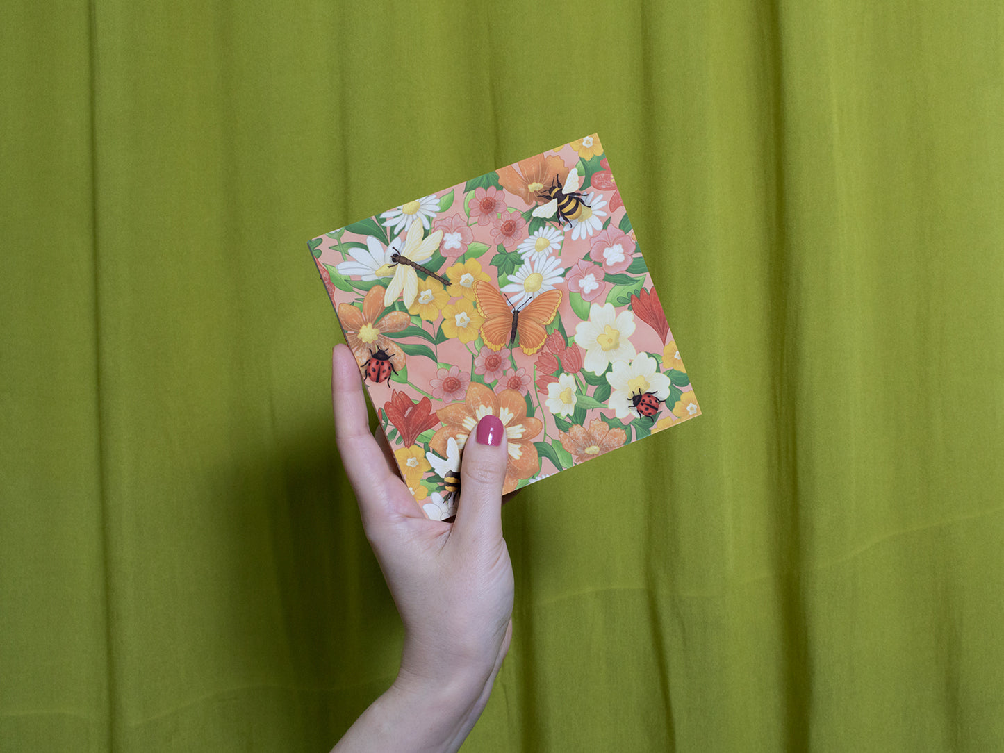 Hand holding a floral square sketchbook with butterflies against a green curtain background