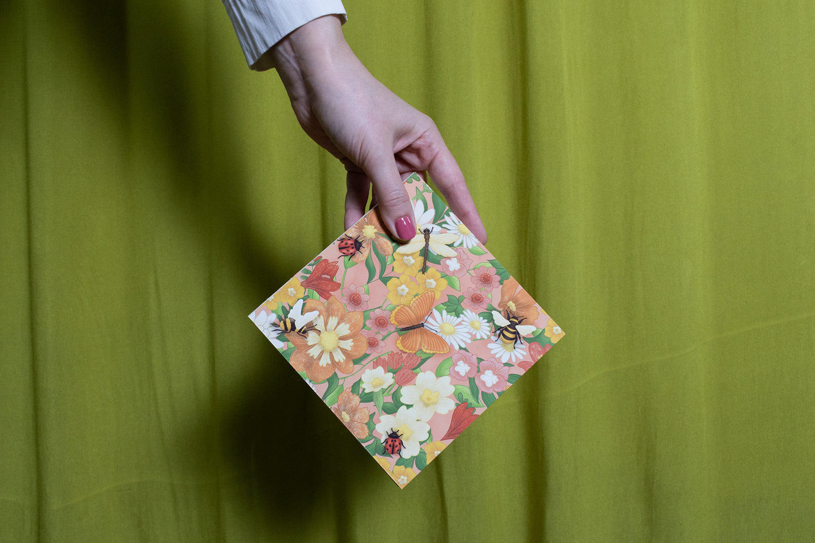 Hand holding a floral square sketchbook against a green curtain background
