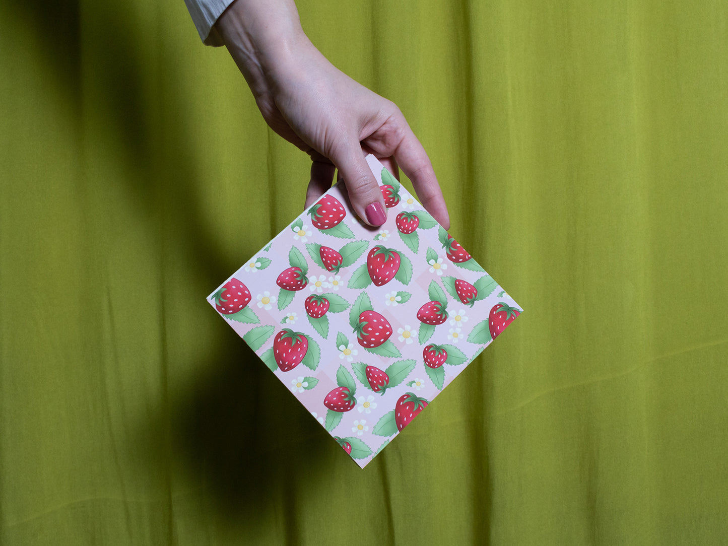 Hand holding a square sketchbook with strawberry pattern against a green background