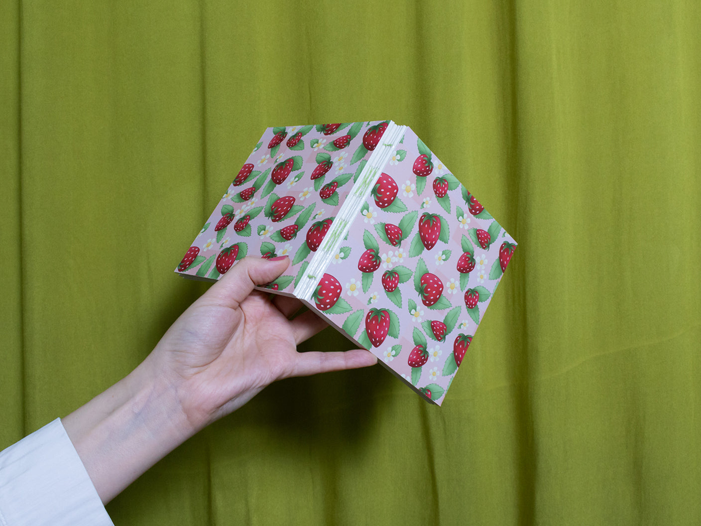 Hand holding a sketchbook with strawberry pattern against a green curtain background